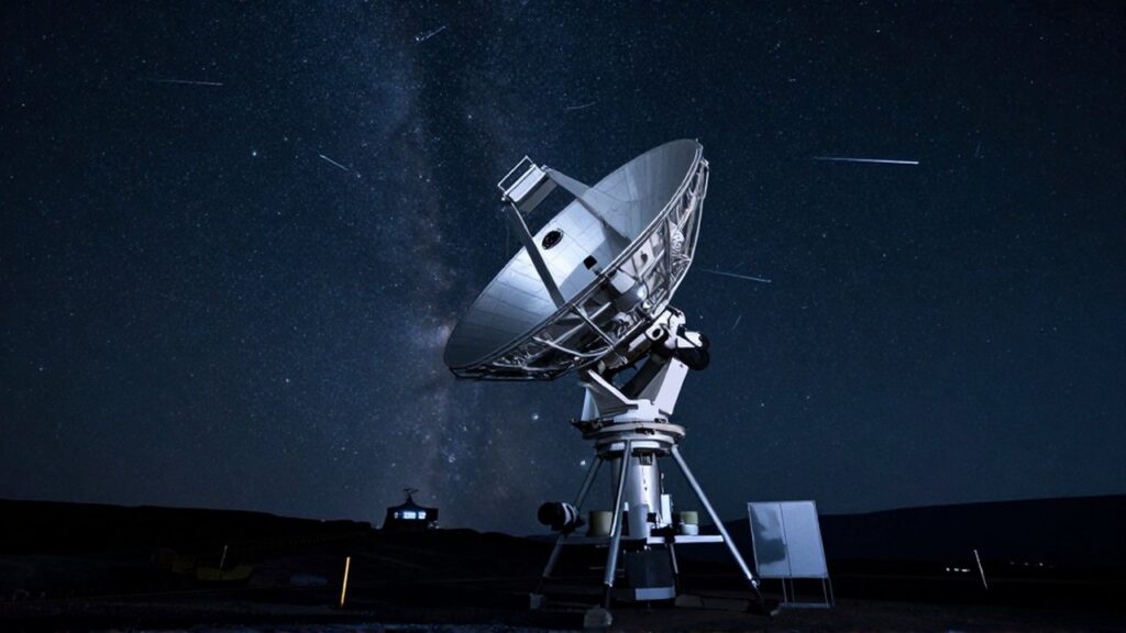 Telescopio de alta tecnología apuntando hacia un cielo nocturno estrellado con meteoros sobre una montaña desolada.